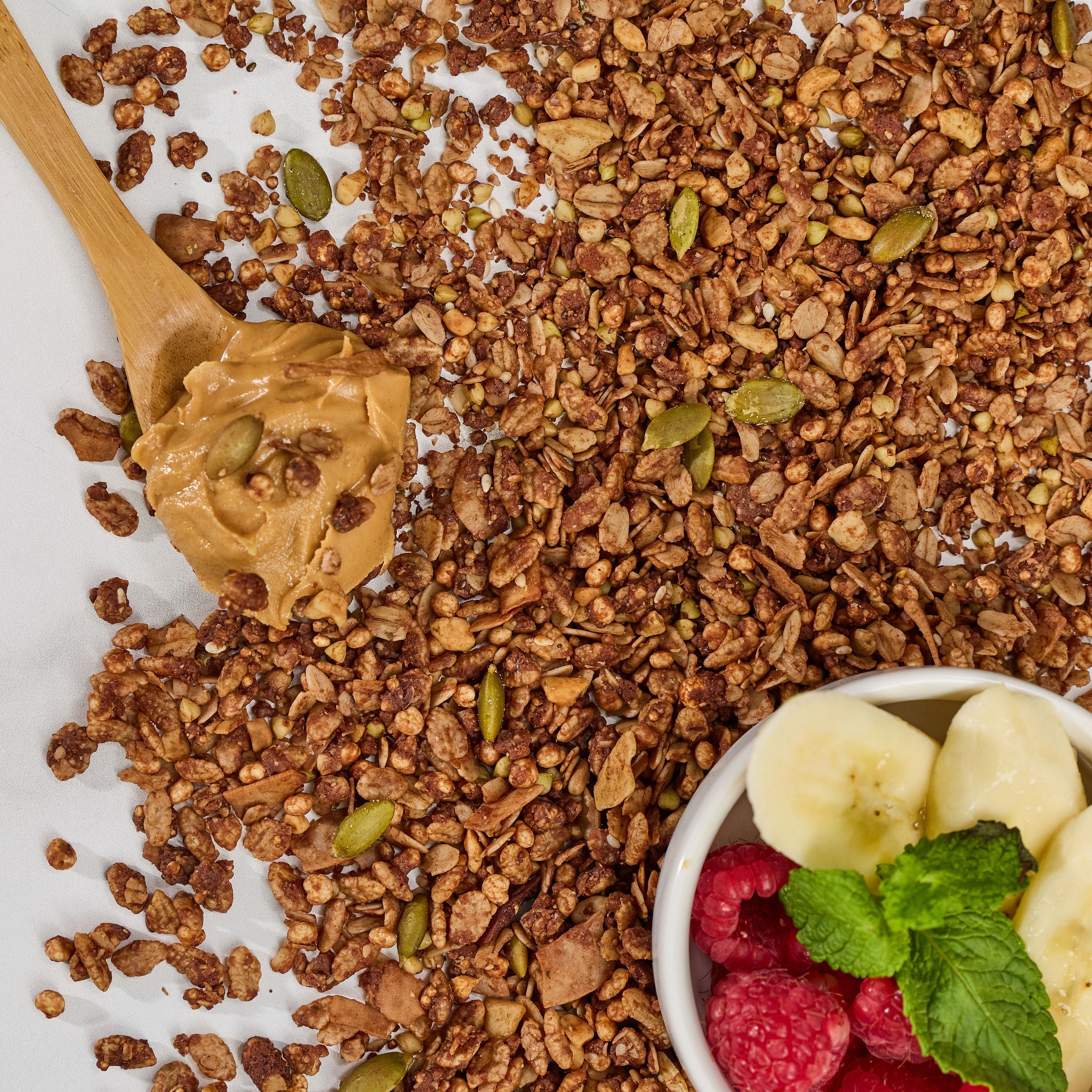 Chocolate and peanut butter granola pieces containing lion's mane extract with peanut butter on a wooden spoon and bowl of raspberries and bananas.