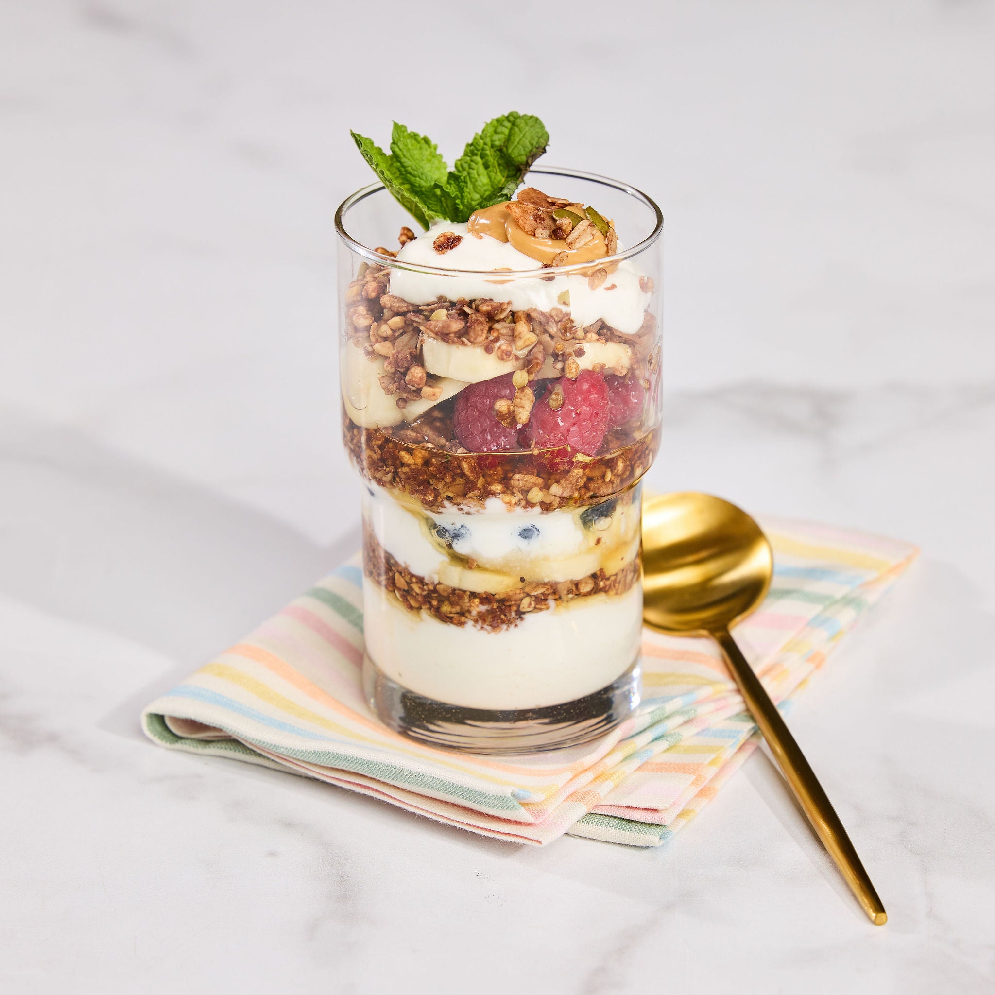 Glass with layers of yogurt, superfood granola, berries, and a dollop of peanut butter on top of a white table.