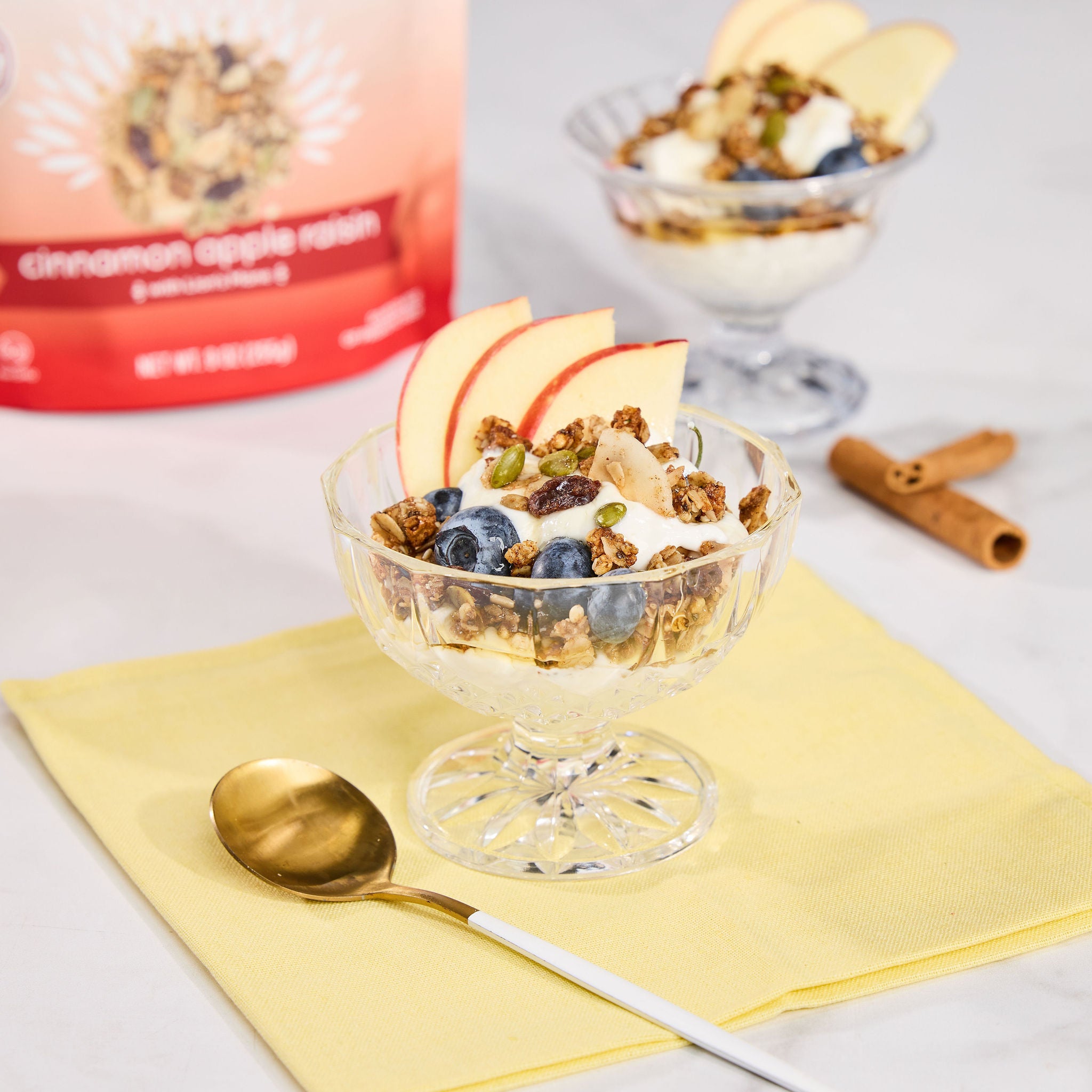 Warm Cinnamon Apple Raisin granola, formulated for gut health served with fresh fruit and yogurt in a glass bowl on a yellow napkin with a spoon.