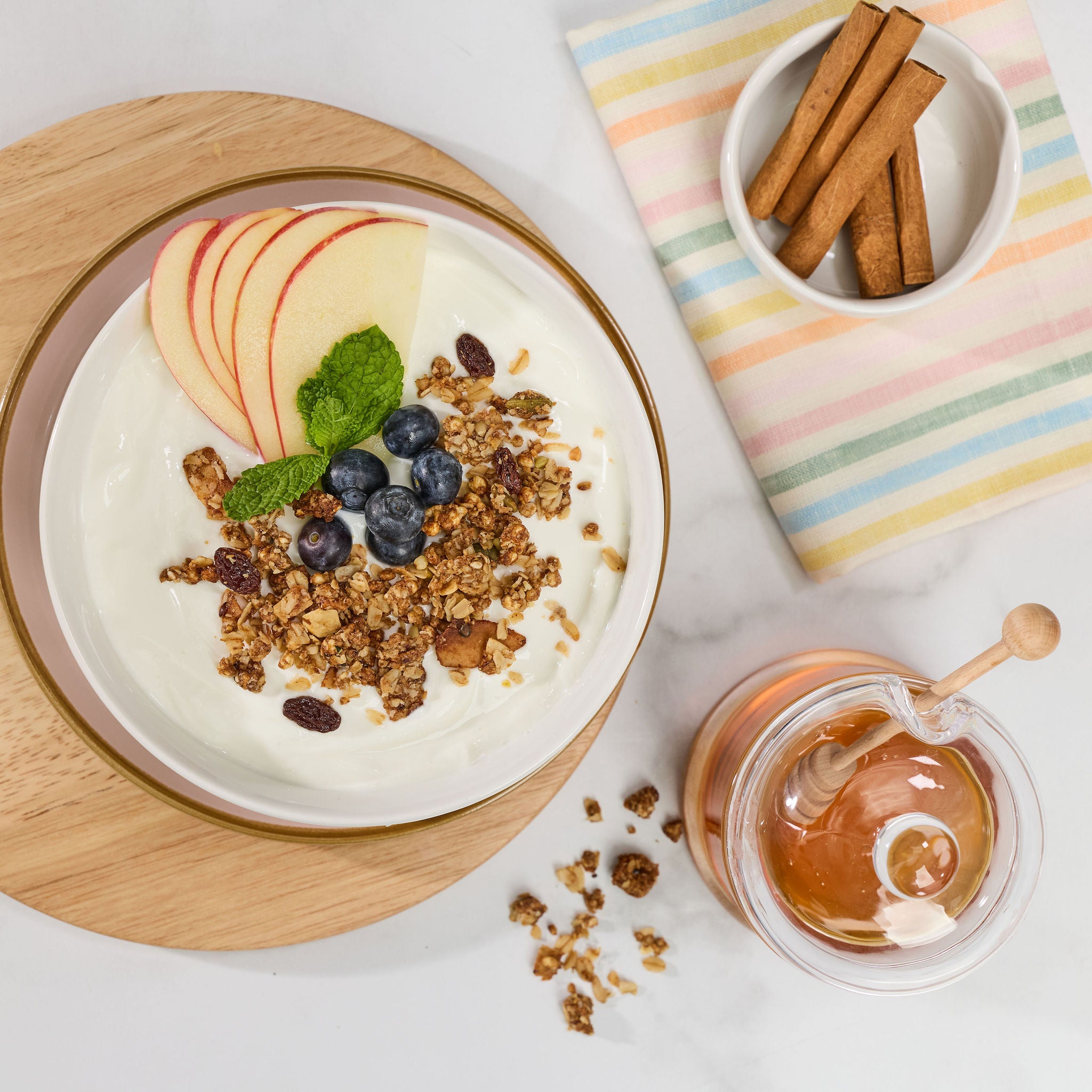 Looking down on a yogurt bowl with granola, sliced apples, blueberries, and cinnamon served on a wooden board with cinnamon sticks and a honey pot on a white table.