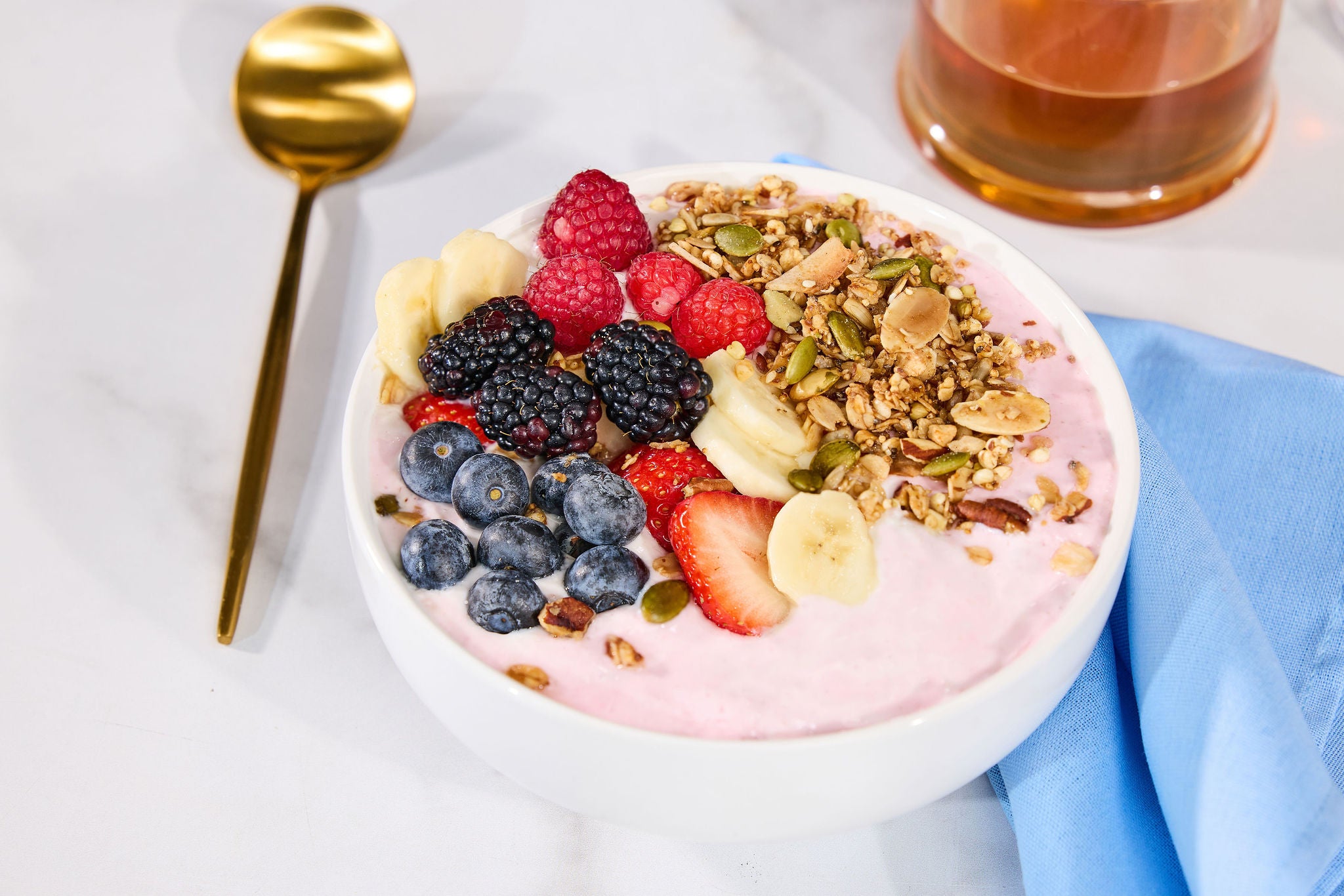 Yogurt bowl with delicious gutBFF granola with lion's mane with fruits on a white table with a blue napkin and gold spoon.